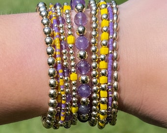 May include: A stack of bracelets with gold beads and purple and yellow accents. The bracelets are worn on a person's wrist.