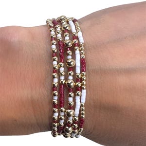 May include: A stack of beaded bracelets in shades of gold, white, and burgundy. The bracelets feature small beads and larger gold-colored accent beads. The bracelets are worn on a wrist.