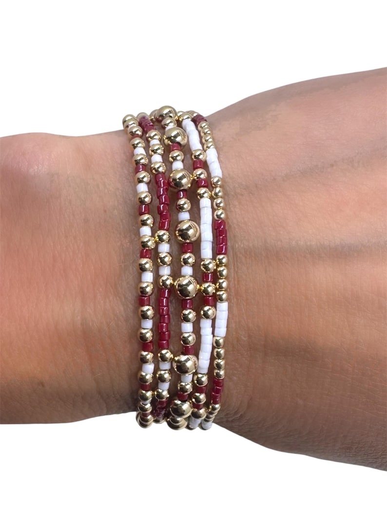 May include: A stack of beaded bracelets in shades of gold, white, and burgundy. The bracelets feature small beads and larger gold-colored accent beads. The bracelets are worn on a wrist.