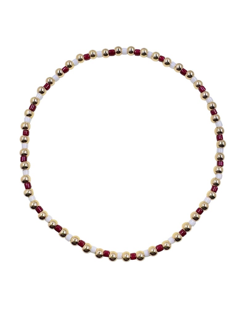 May include: A beaded bracelet featuring a circular design. The bracelet is composed of small, gold-colored, round beads interspersed with white and burgundy beads. The beads are strung together, creating a flexible and stylish accessory.
