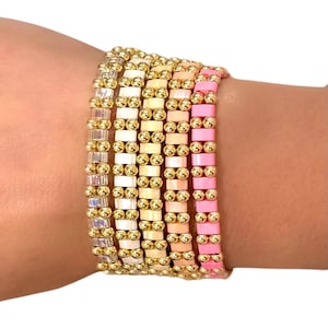 May include: A collection of beaded bracelets in various colors, including gold, white, peach, and pink. The bracelets are made with small, rectangular beads and gold-colored accents. The bracelets are stacked on a wrist.