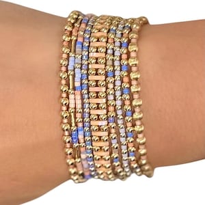 May include: A stack of beaded bracelets in shades of gold, peach, blue, and white. The bracelets feature a mix of small beads and larger, textured gold beads. The bracelets are stacked together, creating a layered look.