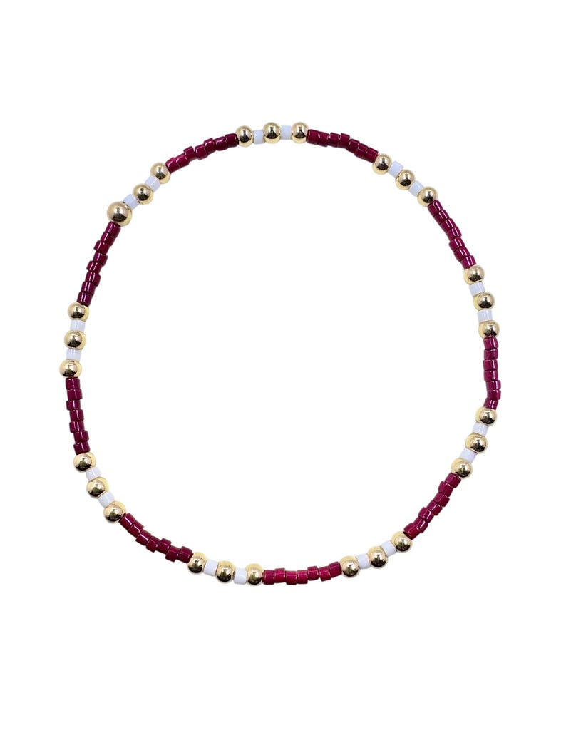May include: A beaded bracelet featuring a circular design. The bracelet is composed of small, burgundy-colored beads, interspersed with gold and white beads. The beads are strung together, creating a flexible and stylish accessory.