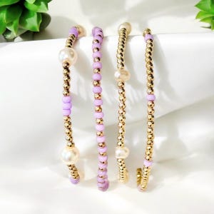 May include: Three delicate bracelets with gold beads and small white pearls. The bracelets are layered and feature lavender colored beads.