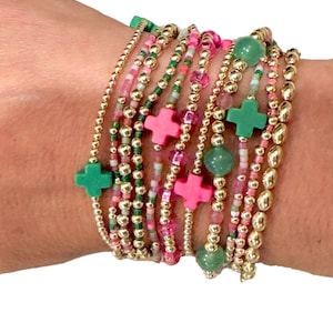 May include: A stack of bracelets with gold beads, green and pink cross charms, and green stone beads.