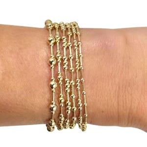 May include: Gold beaded bracelets. The bracelets are made of small, round beads strung together on thin gold chains. The bracelets are stacked on a wrist.