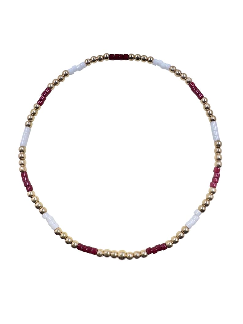 May include: A beaded bracelet featuring a circular design. The bracelet is composed of small, gold-colored beads interspersed with white and burgundy beads. The beads are strung together to form a flexible band.
