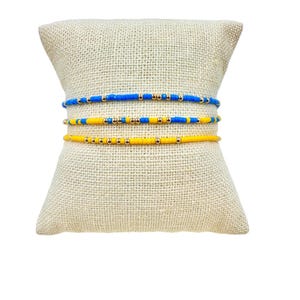 May include: Three beaded bracelets stacked on a beige fabric display. The top bracelet is blue with gold beads, the middle bracelet is yellow with gold beads, and the bottom bracelet is yellow with gold beads.