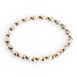 May include: A beaded bracelet featuring gold-colored beads interspersed with white and red beads. The beads are strung together on a red cord, creating a circular design. The bracelet is shown against a white background.
