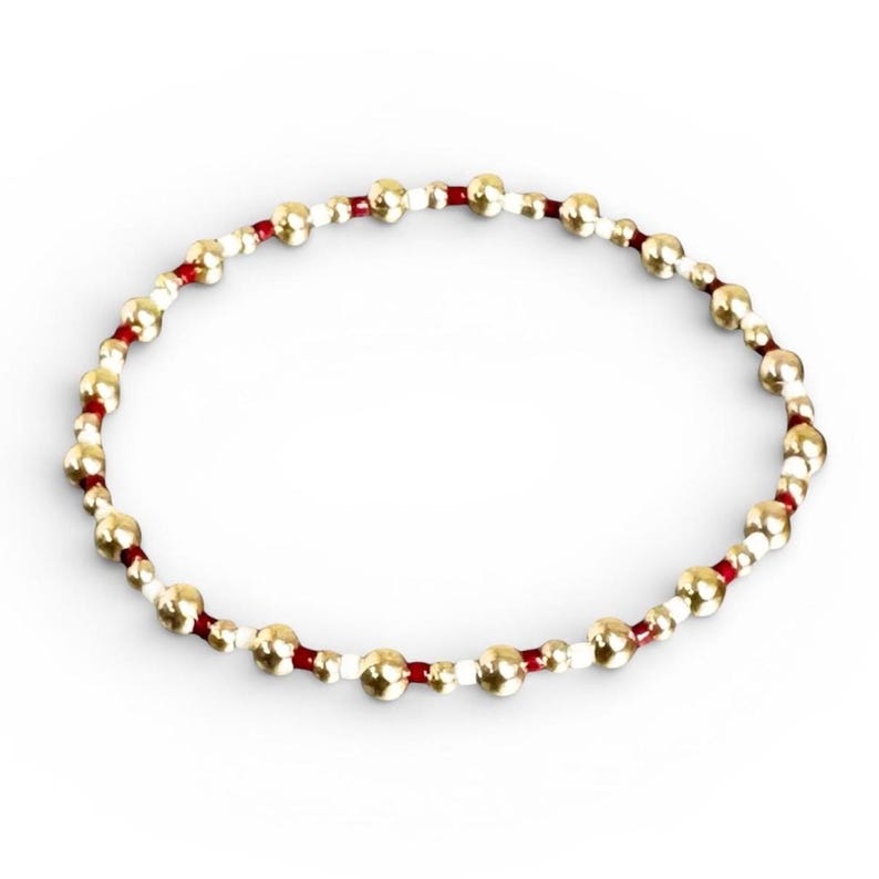 May include: A beaded bracelet featuring gold-colored beads interspersed with white and red beads. The beads are strung together on a red cord, creating a circular design. The bracelet is shown against a white background.