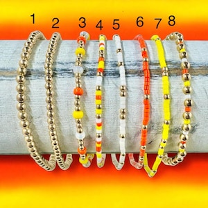 May include: A collection of eight beaded bracelets in various designs and colors. The bracelets include gold, white, orange, yellow, and mixed-color beads. The bracelets are displayed on a light-colored wooden surface against a bright yellow and orange background.