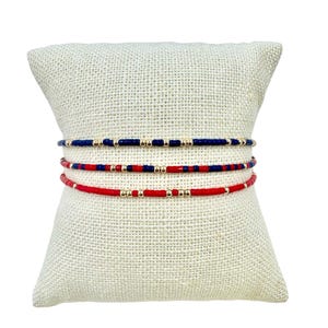 May include: Three beaded bracelets in red, navy blue, and gold, displayed on a textured, off-white pillow. The bracelets feature small, colorful beads interspersed with gold-colored beads. The bracelets are delicate and appear to be handmade.