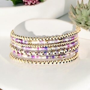 May include: A multi-strand bracelet with gold beads and purple, white, and pink beads. The bracelet is stacked and has a delicate, feminine look.