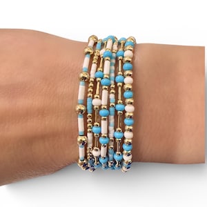 May include: A stack of beaded bracelets with a mix of white, light blue, and gold beads. The bracelets are made of small, round beads and cylindrical gold-colored spacers. The bracelets are worn on a wrist.