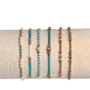 May include: Six beaded bracelets in various designs. The bracelets feature gold beads, turquoise beads, and white pearls. The bracelets are displayed on a cream-colored surface.