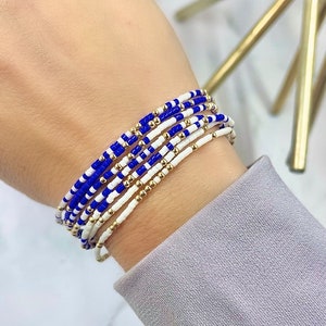 May include: A stack of blue and white beaded bracelets with gold beads. The bracelets are layered on a person's wrist.