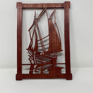 May include: A wooden wall hanging with a cutout design of a sailboat. The sailboat is in the center of the frame and is surrounded by a wooden frame.