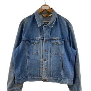 May include: A blue denim jacket with a button closure and a Rustler logo on the pocket. The jacket has a worn look and is hanging on a wooden hanger.
