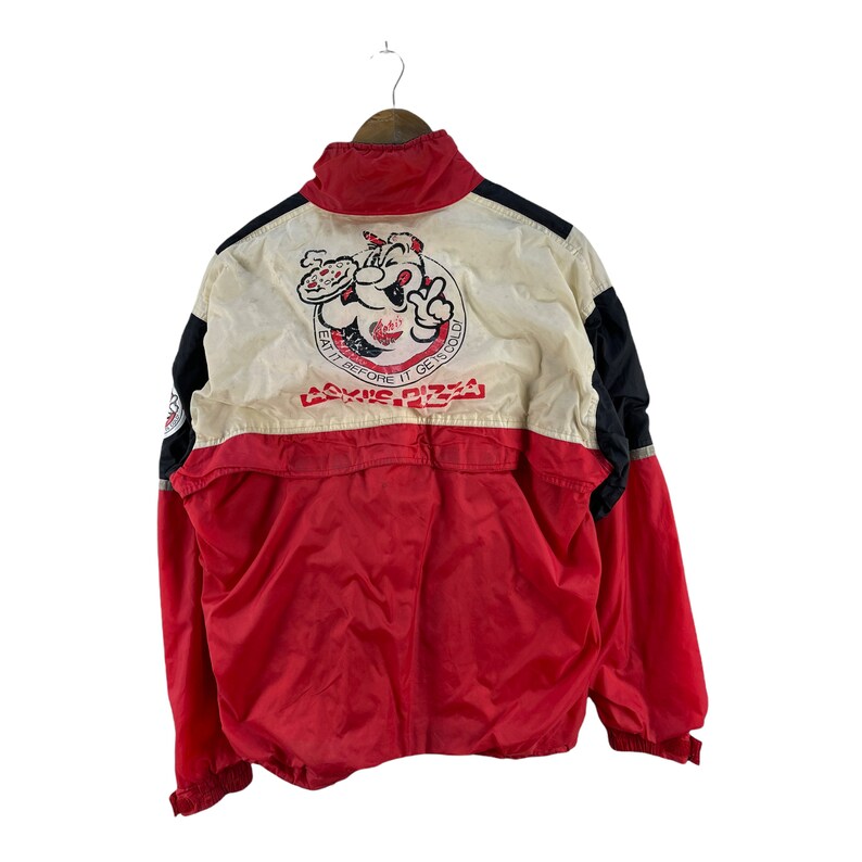 Puede incluir: Una chaqueta cortavientos roja, blanca y negra con un personaje de dibujos animados y el texto "Eat it before it gets cold" y "Cookie Pizza" en la espalda.