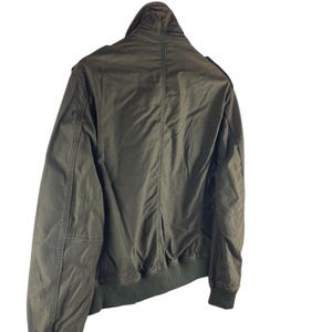 May include: Olive green bomber jacket with a ribbed collar, cuffs, and hem. The jacket has a zip-up front and a stand-up collar. The back of the jacket is visible, showing the seams and the overall shape. A classic outerwear piece.