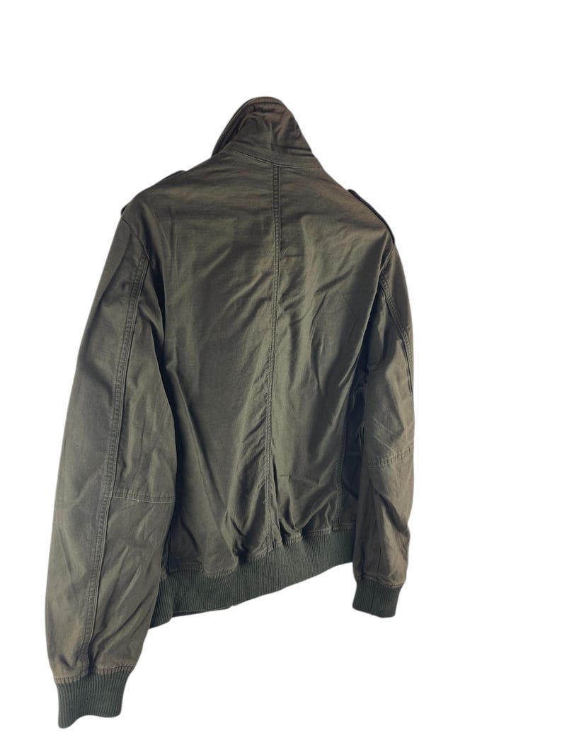 May include: Olive green bomber jacket with a ribbed collar, cuffs, and hem. The jacket has a zip-up front and a stand-up collar. The back of the jacket is visible, showing the seams and the overall shape. A classic outerwear piece.