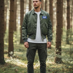 May include: An olive green bomber jacket, white t-shirt, and olive green cargo trousers. The jacket features a small patch on the chest. The person is in a forest, surrounded by trees and green foliage.