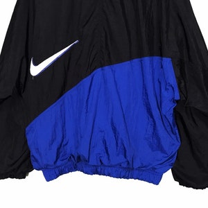 Op de afbeelding: Een zwart en blauw windjack met een wit Nike swoosh-logo aan de linkerkant van de borst.