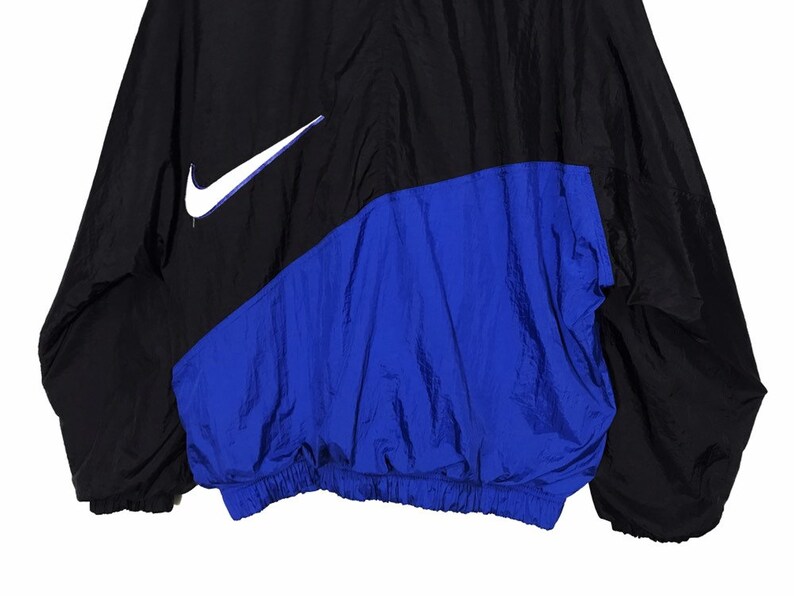 Op de afbeelding: Een zwart en blauw windjack met een wit Nike swoosh-logo aan de linkerkant van de borst.