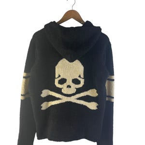 May include: Black hooded sweater with a skull and crossbones design on the back. The sweater has gold stripes on the sleeves and is hanging on a wooden hanger. The skull and crossbones are in a cream color.