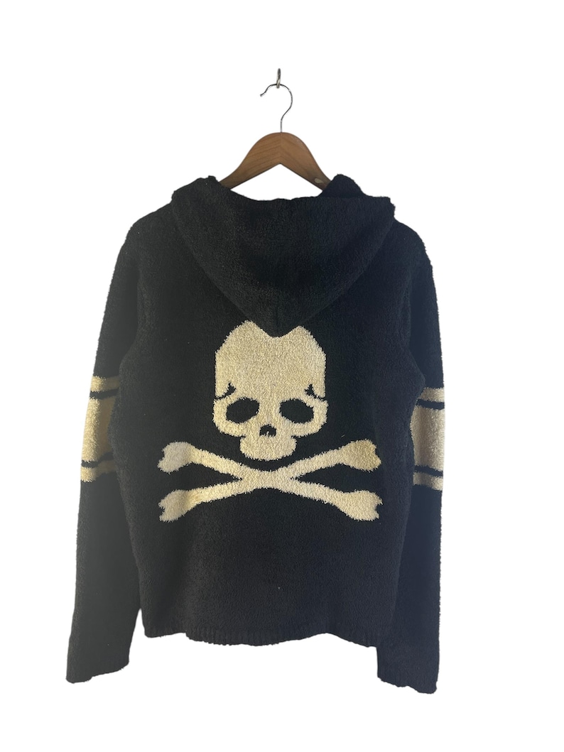 May include: Black hooded sweater with a skull and crossbones design on the back. The sweater has gold stripes on the sleeves and is hanging on a wooden hanger. The skull and crossbones are in a cream color.