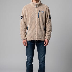 May include: A tan fleece jacket with a high collar and a full zipper, paired with dark blue jeans. The jacket features navy blue accents on the pockets and a logo on the chest. The model is wearing white sneakers.