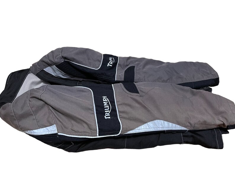 Puede incluir: Una chaqueta de moto gris y negra con la palabra "TRIUMPH" impresa en el lateral.