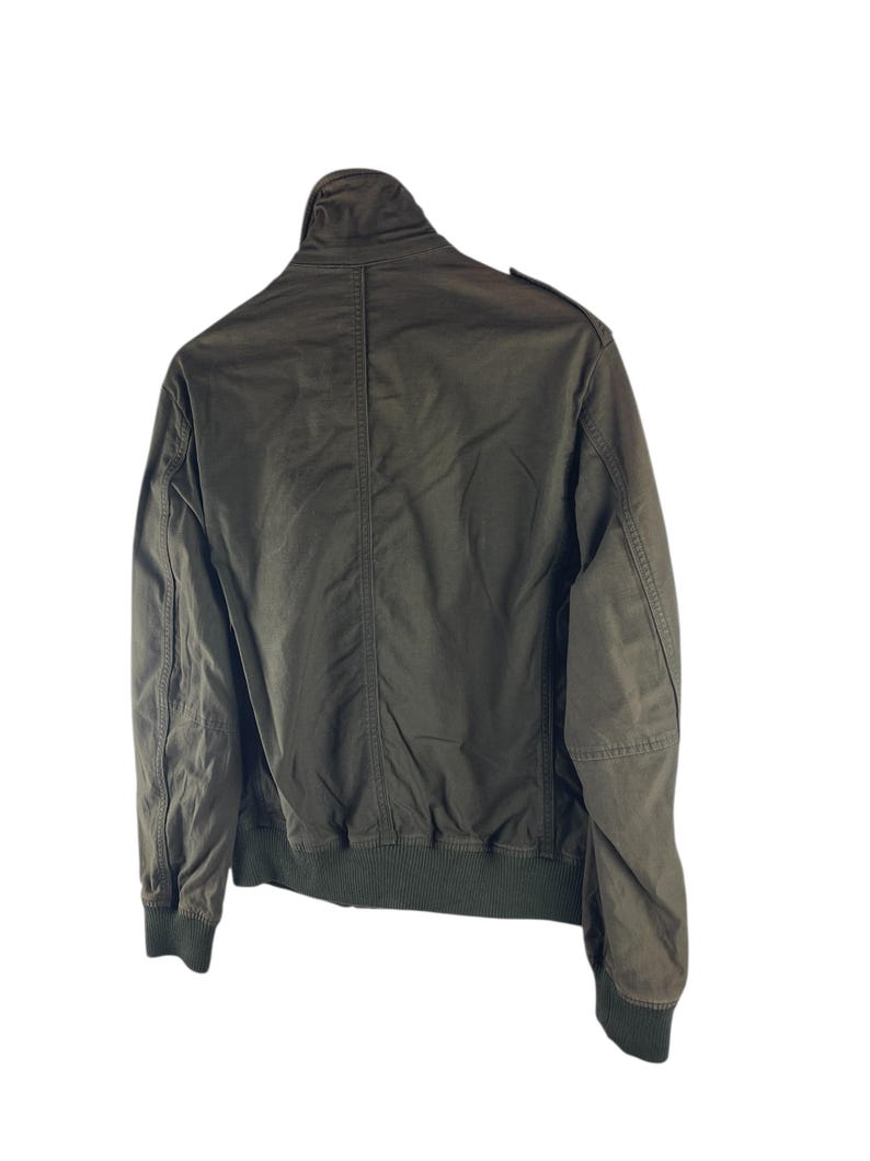 May include: Olive green bomber jacket with a ribbed collar, cuffs, and hem. The jacket has a full-length zipper and two front pockets. The back of the jacket is visible, showing the seams and construction details. A classic outerwear piece.