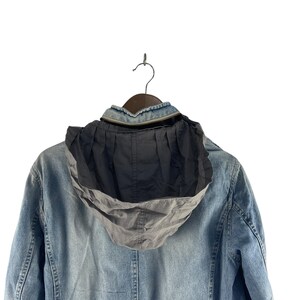 Puede incluir: Chaqueta vaquera azul claro con capucha gris, colgada de una percha marr&oacute;n. La chaqueta tiene cierre de cremallera y forro de capucha plisado. La tela vaquera muestra un lavado descolorido, y la chaqueta es una prenda de abrigo informal.