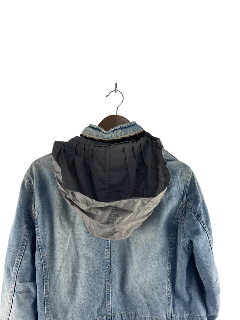 Puede incluir: Chaqueta vaquera azul claro con capucha gris, colgada de una percha marr&oacute;n. La chaqueta tiene cierre de cremallera y forro de capucha plisado. La tela vaquera muestra un lavado descolorido, y la chaqueta es una prenda de abrigo informal.