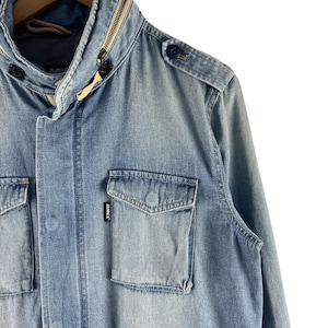 Puede incluir: Chaqueta vaquera azul claro con cuello con cremallera y m&uacute;ltiples bolsillos. La chaqueta tiene una charretera abotonada y una peque&ntilde;a etiqueta de marca en el bolsillo. La chaqueta est&aacute; colgada en una percha de madera.