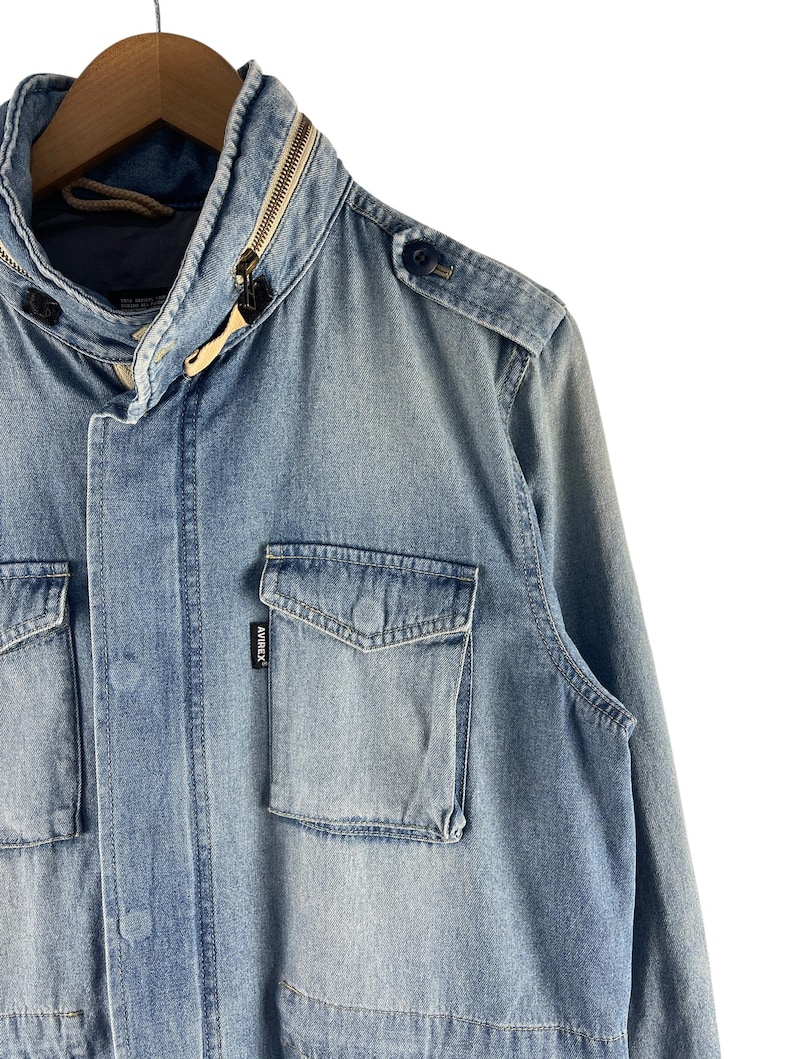 Puede incluir: Chaqueta vaquera azul claro con cuello con cremallera y m&uacute;ltiples bolsillos. La chaqueta tiene una charretera abotonada y una peque&ntilde;a etiqueta de marca en el bolsillo. La chaqueta est&aacute; colgada en una percha de madera.