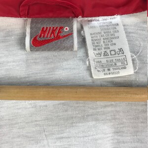 Puede incluir: Una chaqueta cortavientos roja Nike con una etiqueta gris que dice "Nike" y tiene instrucciones de cuidado para lavar y secar la chaqueta. La etiqueta tambi&eacute;n incluye la talla, "KL(18/20)XLB", y el pa&iacute;s de origen, "Hecho en Tailandia".