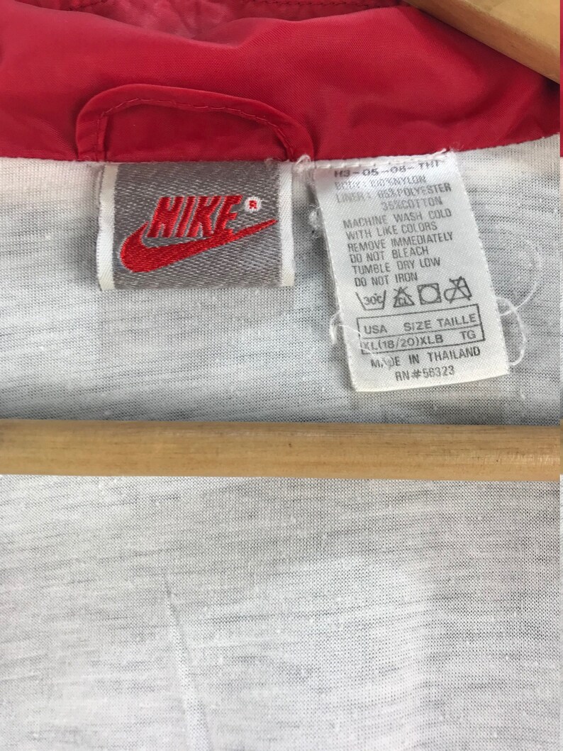 Puede incluir: Una chaqueta cortavientos roja Nike con una etiqueta gris que dice "Nike" y tiene instrucciones de cuidado para lavar y secar la chaqueta. La etiqueta tambi&eacute;n incluye la talla, "KL(18/20)XLB", y el pa&iacute;s de origen, "Hecho en Tailandia".