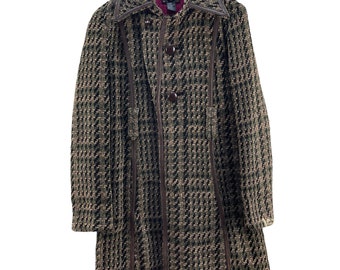 May include: A long, tweed coat with a brown, black, and tan houndstooth pattern. The coat features brown leather trim along the front and collar, with brown buttons. The coat is hanging on a wooden hanger.