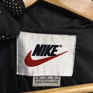 Op de afbeelding: Een wit label met het woord "NIKE" in zwart met een rood swoosh-logo. Het label vermeldt ook "MADE IN THAILAND" en heeft drie "M" voor de maat.