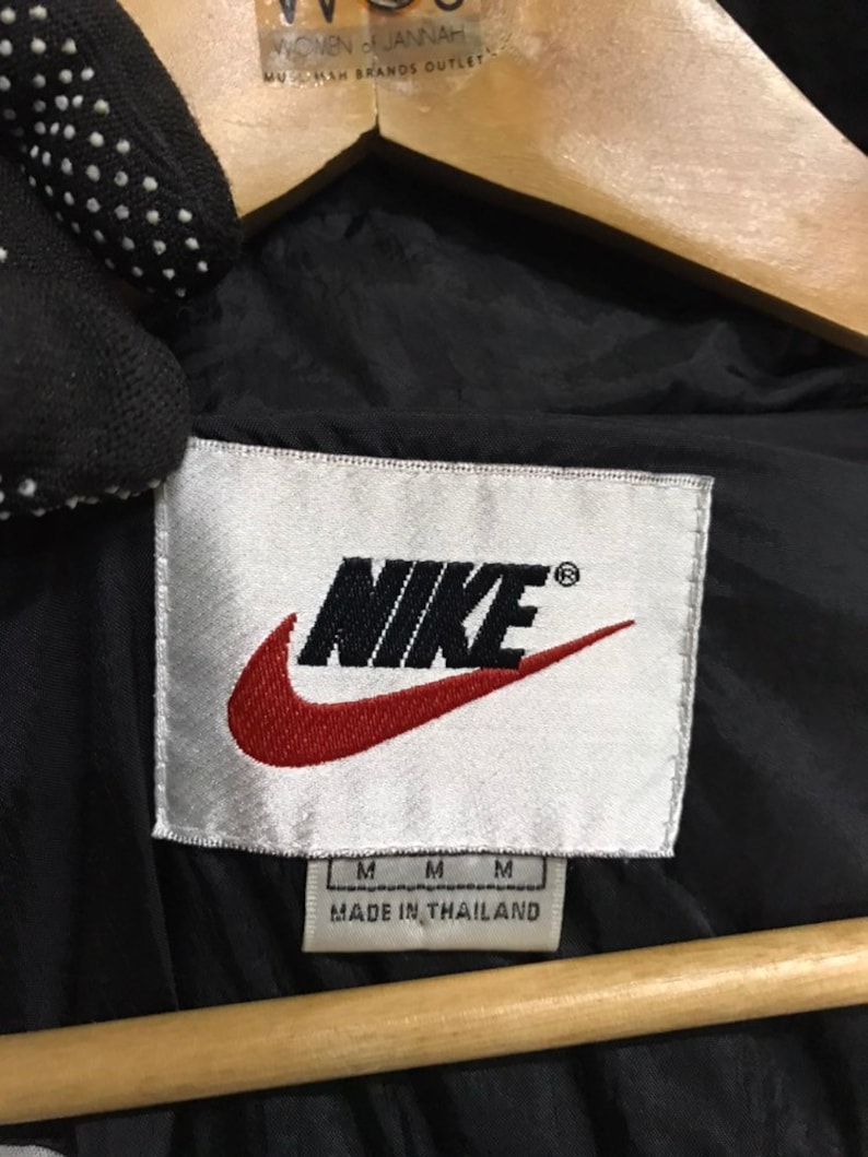 Op de afbeelding: Een wit label met het woord "NIKE" in zwart met een rood swoosh-logo. Het label vermeldt ook "MADE IN THAILAND" en heeft drie "M" voor de maat.