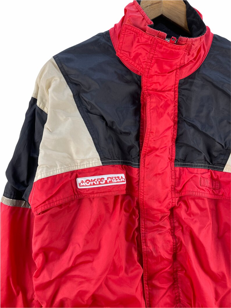 Puede incluir: Una chaqueta roja, negra y blanca con cierre de cremallera y un logotipo de "Aoki's Pizza" en la parte delantera.