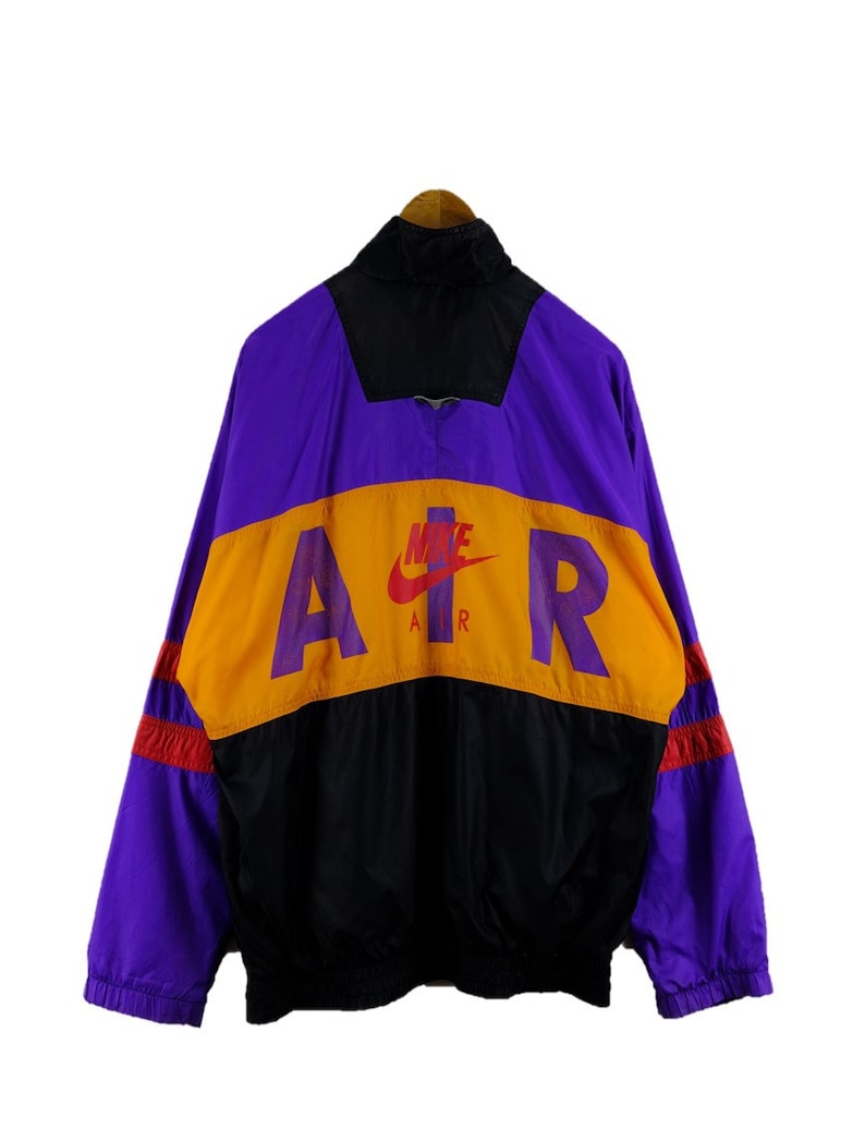 Puede incluir: Una chaqueta cortavientos de color morado, negro y naranja con la palabra "AIR" en letras grandes en la espalda. El logotipo de Nike swoosh est&aacute; por encima de la palabra "AIR" con la palabra "AIR" escrita debajo del swoosh.