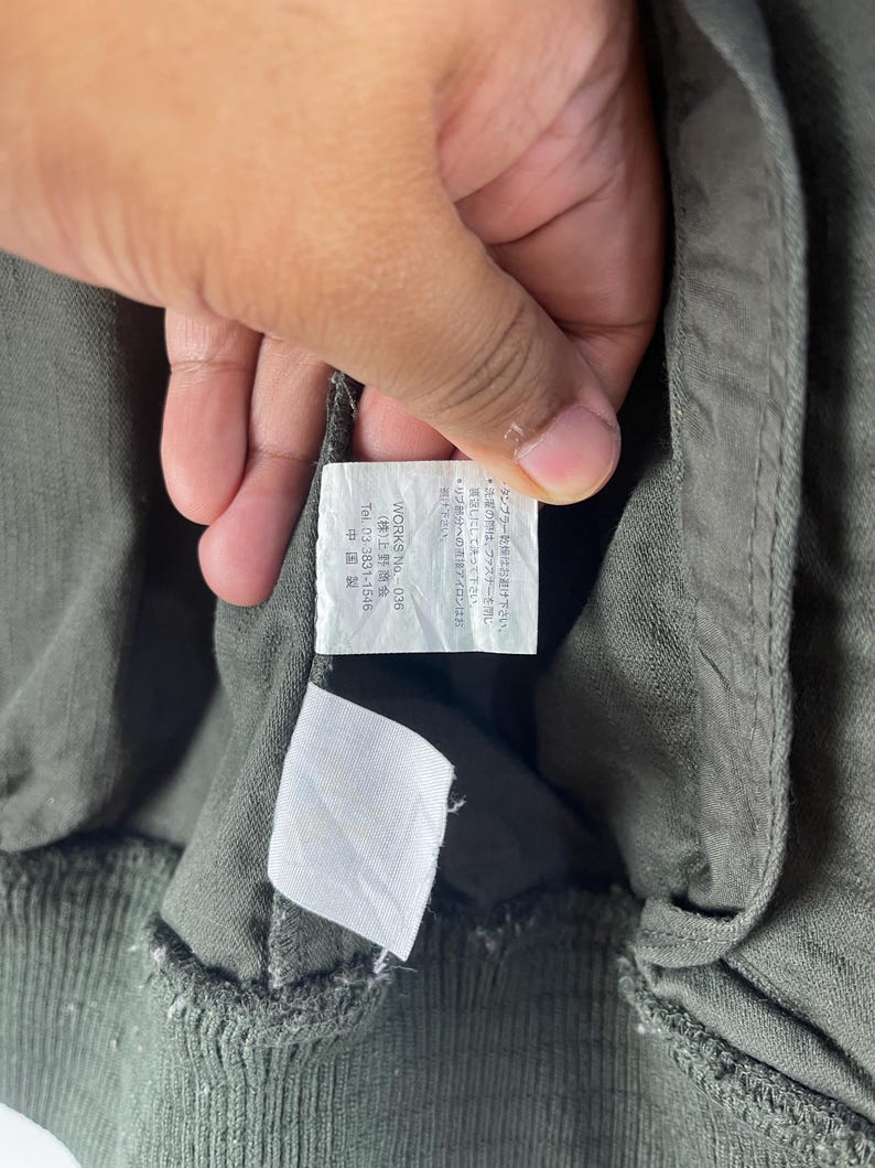 May include: Close-up of a green garment with a tag held by a hand. The tag has Japanese text and the words "WORKS No. 036" and "Tel. 03-3831-1546". The garment is made in China.