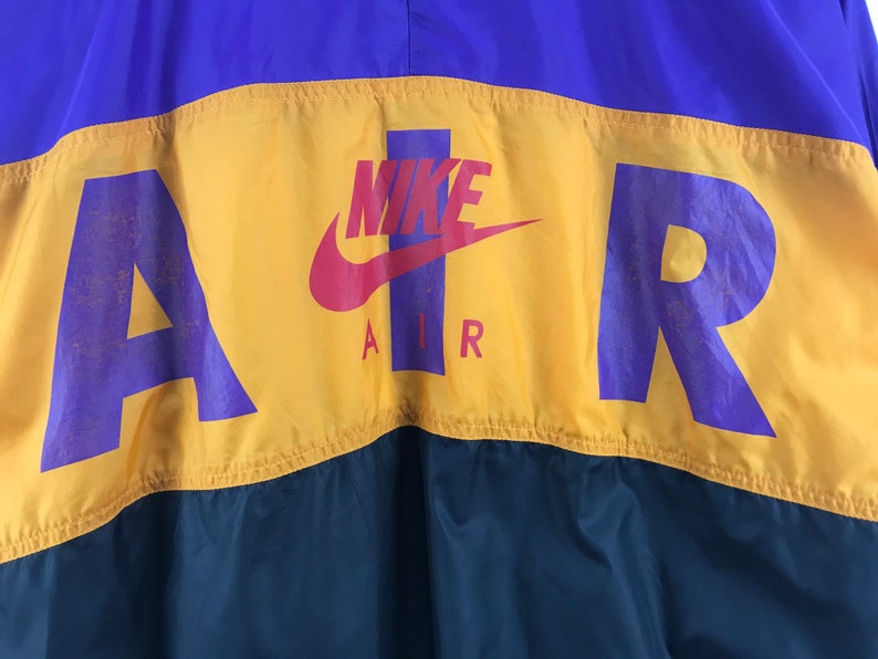 Puede incluir: Una chaqueta cortavientos amarilla, morada y verde con la palabra "AIR" en letras moradas y un logotipo Nike swoosh en rojo. La palabra "NIKE" est&aacute; escrita en letras blancas sobre el swoosh.