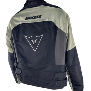 K&ouml;nnte beinhalten: Schwarze und graue Motorradjacke mit dem Markennamen "DAINESE" &uuml;ber den Schultern. Die Jacke hat ein wei&szlig;es Logo auf dem R&uuml;cken und verstellbare Riemen an der Taille. Die Jacke h&auml;ngt an einem Holzkleiderb&uuml;gel.