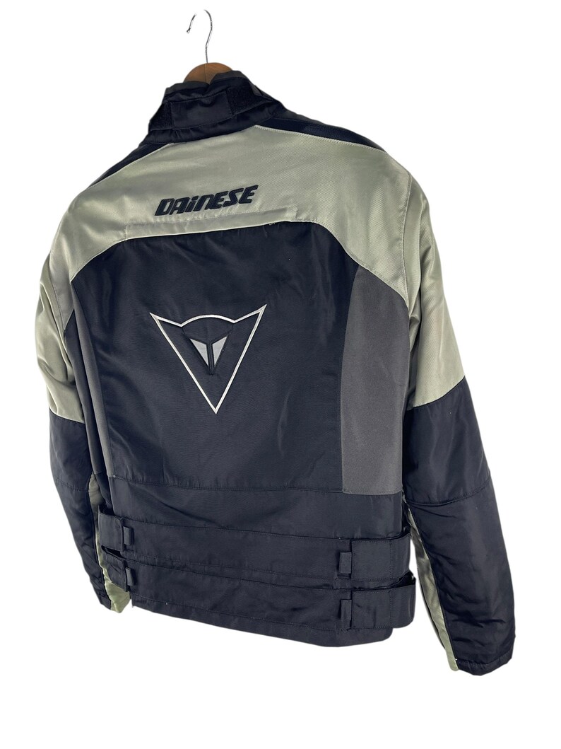 K&ouml;nnte beinhalten: Schwarze und graue Motorradjacke mit dem Markennamen "DAINESE" &uuml;ber den Schultern. Die Jacke hat ein wei&szlig;es Logo auf dem R&uuml;cken und verstellbare Riemen an der Taille. Die Jacke h&auml;ngt an einem Holzkleiderb&uuml;gel.