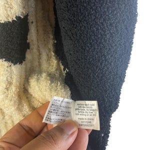May include: Close-up of a black and cream-colored plush fabric. A tag with washing instructions in English and Japanese is held in the frame. The tag reads "machine wash cold with like colors, gentle cycle, no bleach, tumble dry low/cool setting or air dry."