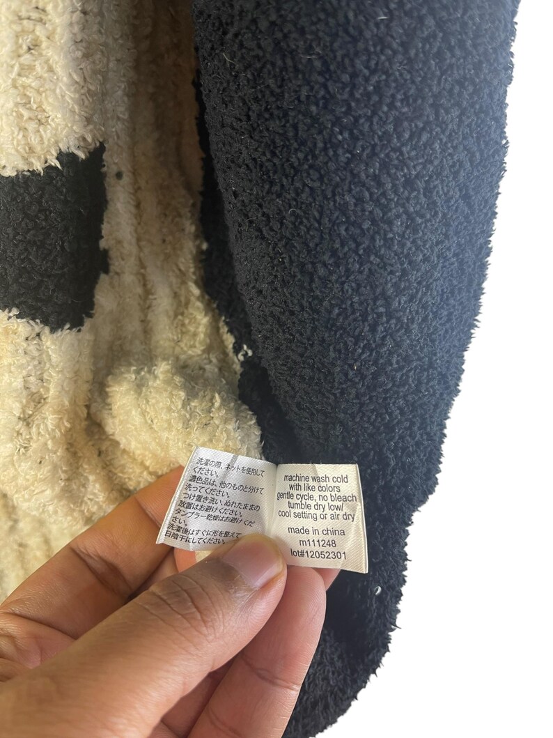 May include: Close-up of a black and cream-colored plush fabric. A tag with washing instructions in English and Japanese is held in the frame. The tag reads "machine wash cold with like colors, gentle cycle, no bleach, tumble dry low/cool setting or air dry."